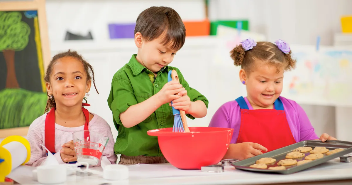 Three young children engage in literacy-rich dramatic play as they bake together in a kitchen. One whisks in a red bowl, another holds a tray of cookies, and the third measures ingredients—all smiling and wearing aprons.