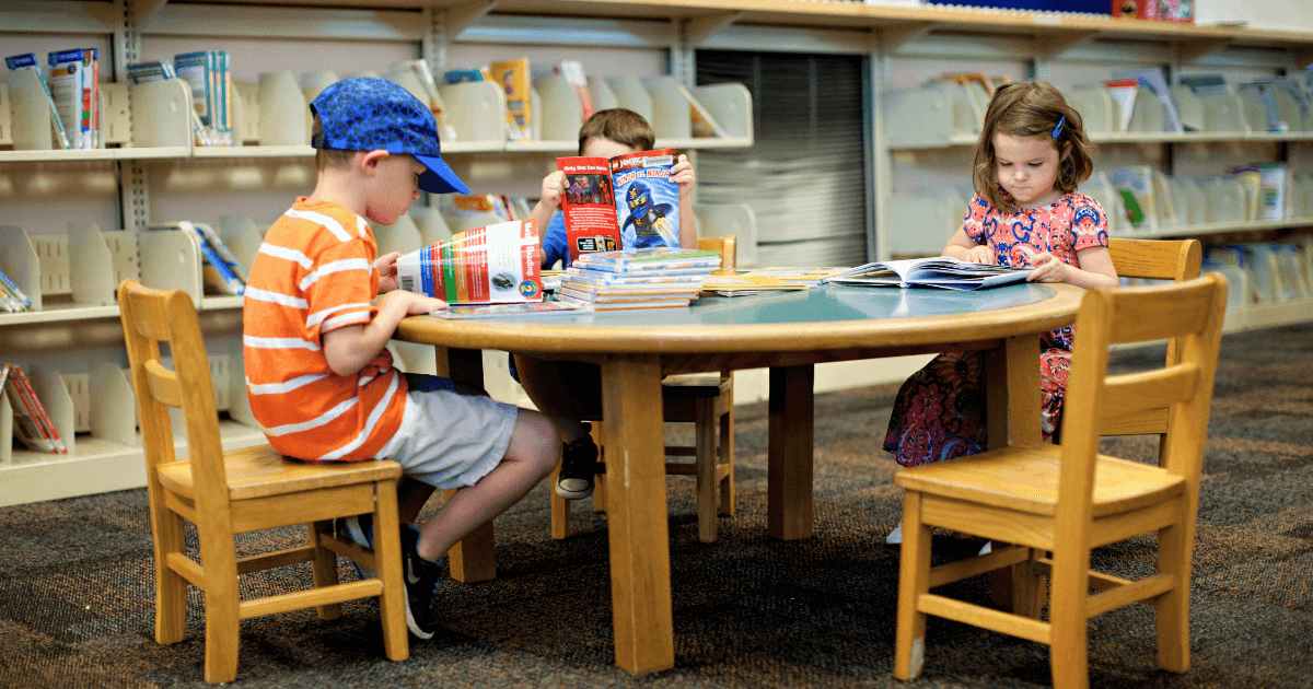 Three young children sit at a round table in a library, each reading books. Shelves of books line the background, reflecting how local business collaborations help create vibrant, welcoming spaces for young readers to learn and grow.