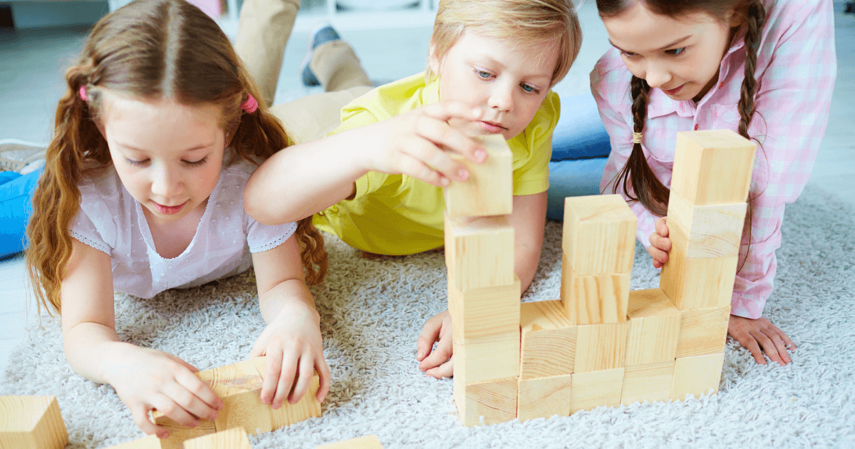 Three young children are lying on a carpet, building a structure together with wooden blocks. They are focused and engaged in creative loose parts play, using their imagination to create and collaborate.