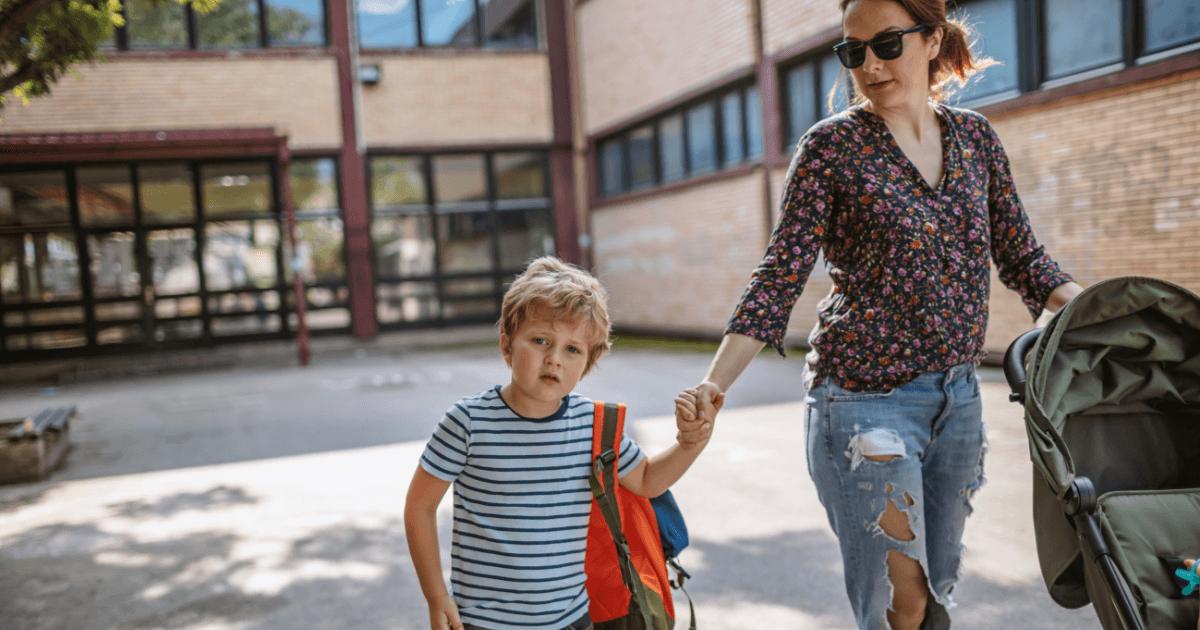 A woman wearing sunglasses and ripped jeans holds the hand of a young boy with a striped shirt and backpack while pushing a stroller outside a school building, possibly arriving for mid-year enrollment.