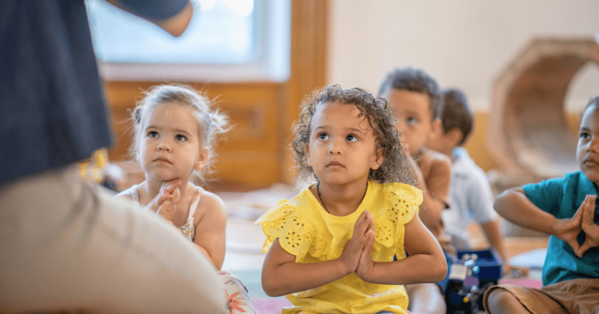 Young children sit on the floor, hands pressed together in a prayer-like gesture, practicing mindfulness as they attentively look toward an adult out of view. The setting appears to be a classroom or daycare.