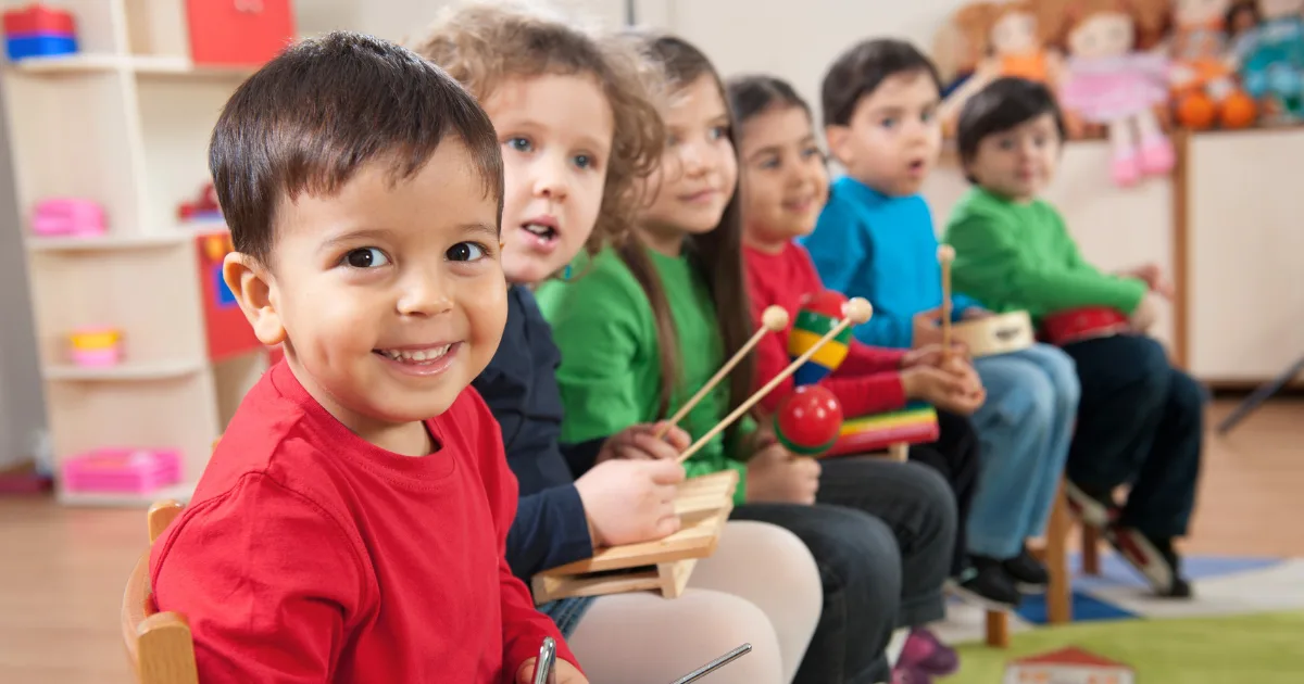 A group of young children in a mixed-age classroom sit in a row, smiling and holding musical instruments. The child in front looks at the camera, while the others are focused on playing their instruments.