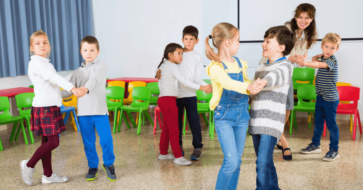 A group of children in a classroom stand in pairs, holding hands as if practicing a dance during a music and movement activity. Brightly colored chairs line the wall, and an adult woman watches and smiles in the background.