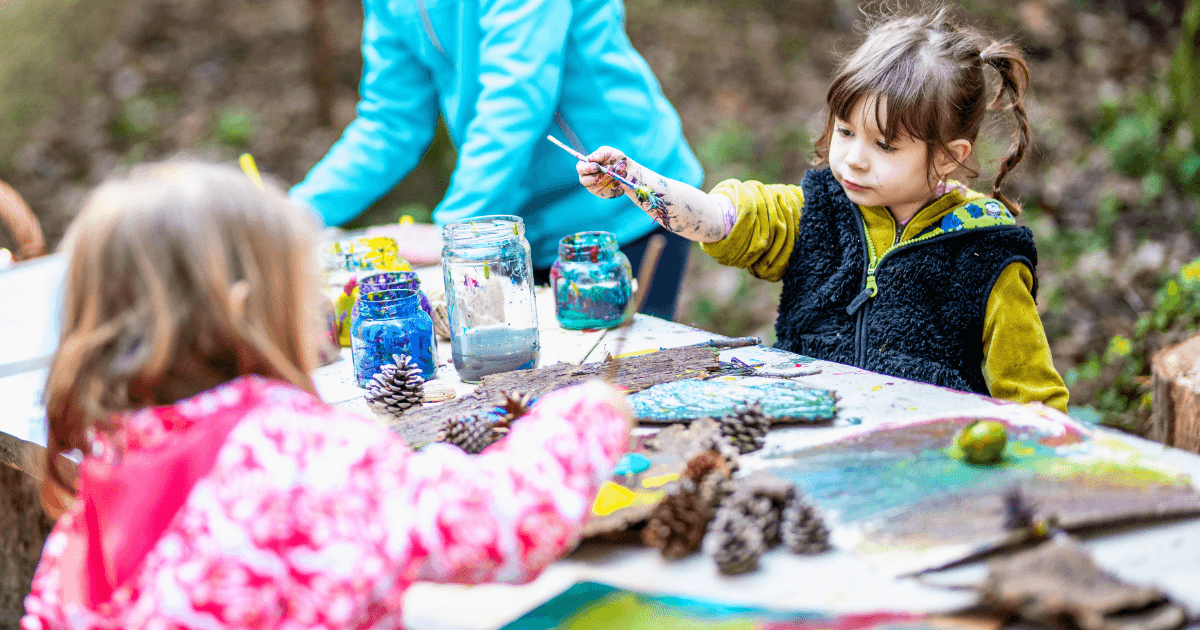 Two young children sit at an outdoor table painting pinecones and wooden boards with bright colors—an engaging activity often found in nature-based preschools, surrounded by art supplies and jars of paint. Trees and greenery fill the background.