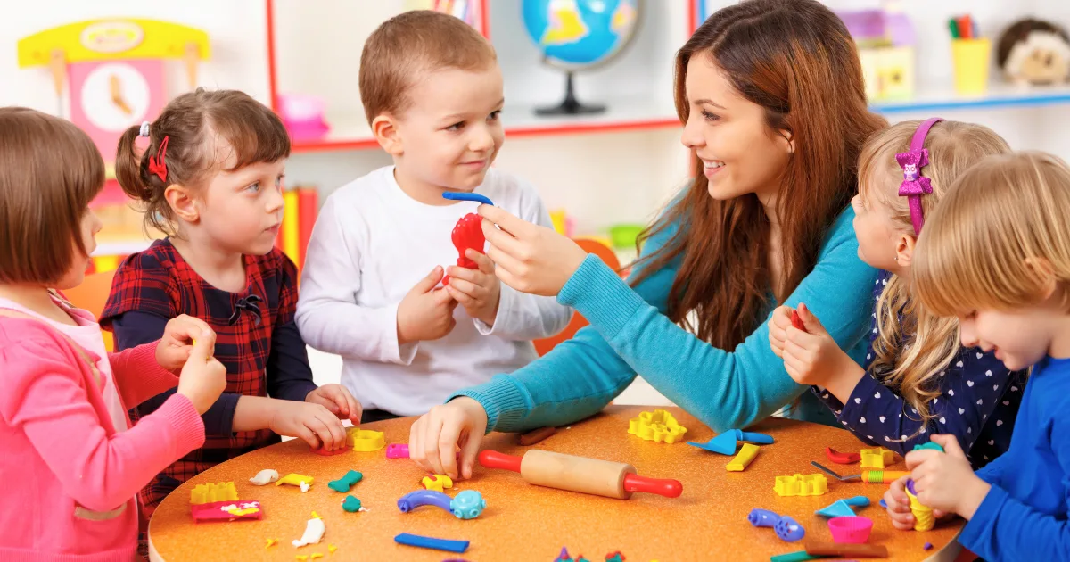 A smiling teacher sits at a table with five young children, playing with colorful clay and tools in a bright classroom. The children are engaged and interacting, creating joyful moments at this vibrant nursery school near me.