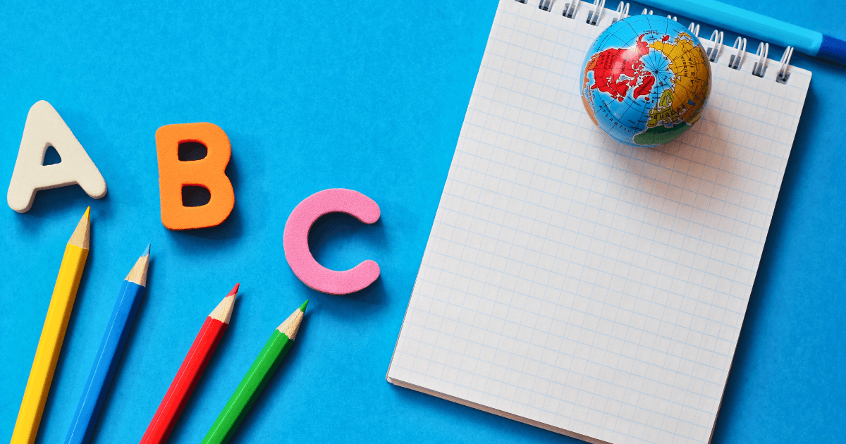 Colorful alphabet letters A, B, and C, four colored pencils, a small globe, and a blank checkered notepad are arranged on a bright blue background reminiscent of the ocean.