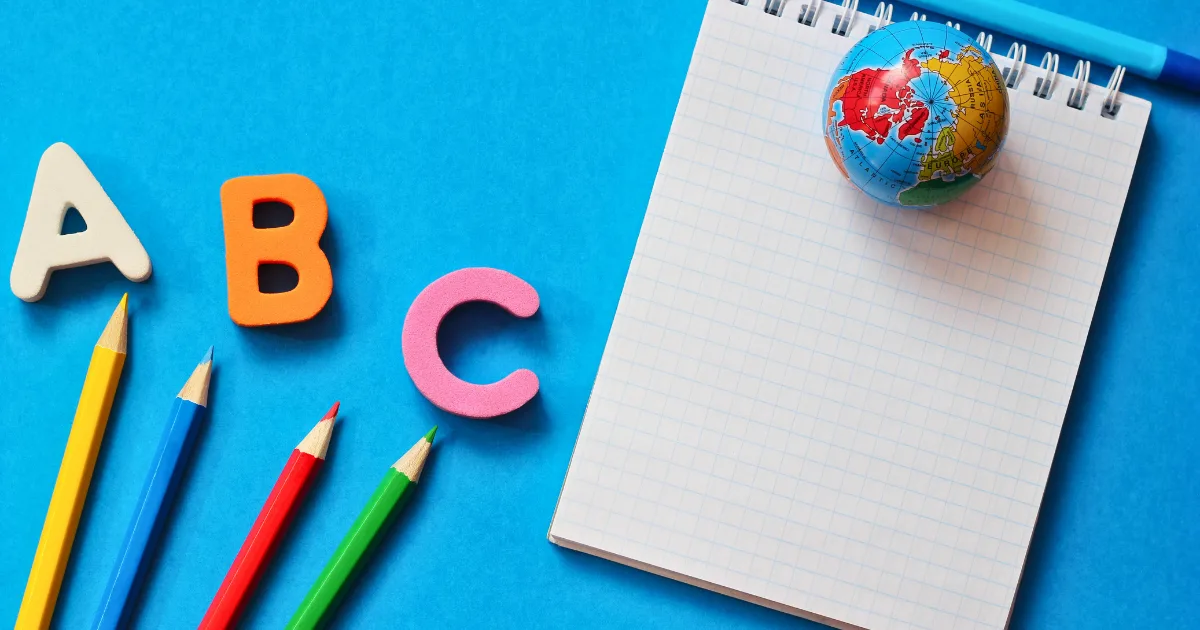 Colorful alphabet letters A, B, and C, four colored pencils, a small globe, and a blank checkered notepad are arranged on a bright blue background reminiscent of the ocean.