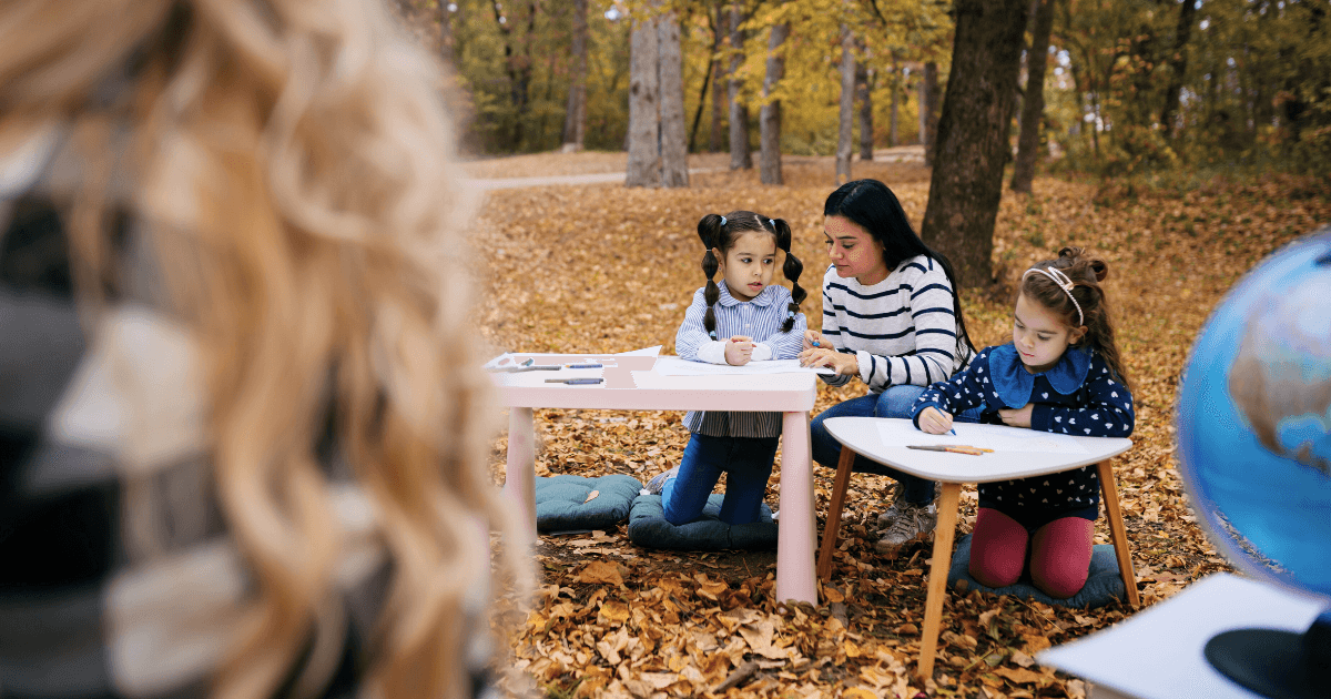 A woman helps two young girls with their schoolwork at small tables in an outdoor classroom set in a forested area covered in autumn leaves. A globe and school supplies sit on the tables, while another person is blurred in the foreground.
