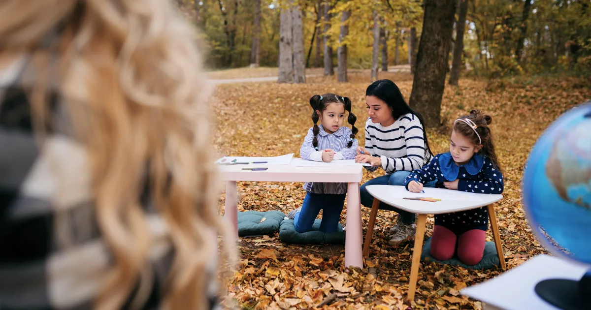 A woman helps two young girls with their schoolwork at small tables in an outdoor classroom set in a forested area covered in autumn leaves. A globe and school supplies sit on the tables, while another person is blurred in the foreground.
