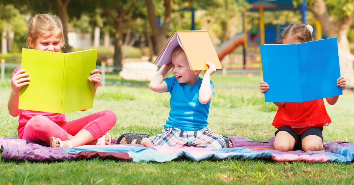 Three children sit on a blanket on grass; two read colorful books, while the child in the middle balances a book on their head. Trees and outdoor learning stations are visible in the background.