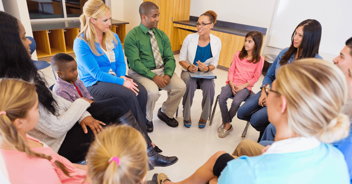 A diverse group of adults and children sit in a circle in a classroom, engaging in a discussion similar to parent-teacher conferences. One woman holds a clipboard and appears to be leading the conversation.