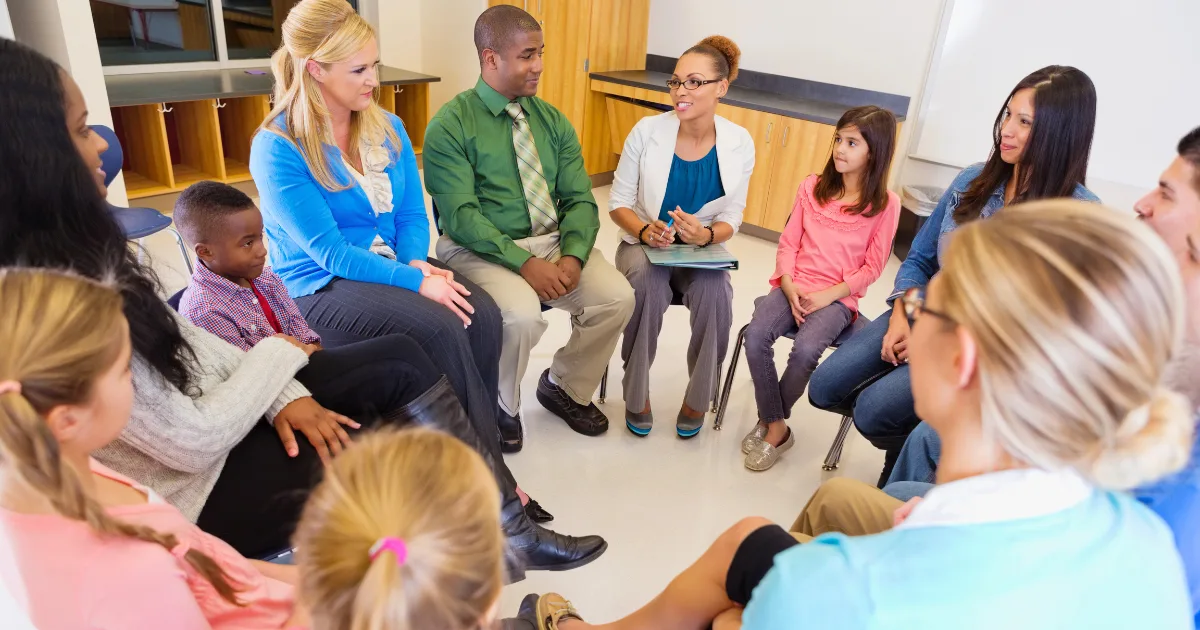 A diverse group of adults and children sit in a circle in a classroom, engaging in a discussion similar to parent-teacher conferences. One woman holds a clipboard and appears to be leading the conversation.
