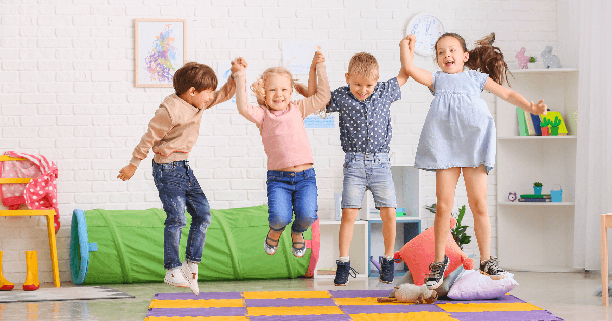 Four young children are joyfully jumping together on colorful foam mats in a bright, cheerful playroom, building physical literacy amid toys and vibrant decorations in the background.