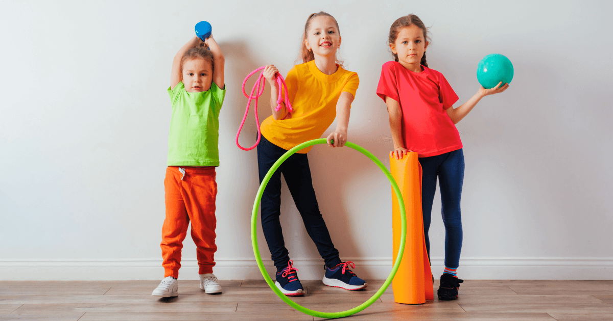 Three children stand indoors against a white wall, each holding sports equipment like a dumbbell, jump rope, and hula hoop—tools that support movement and cognitive development. They wear colorful t-shirts and pants, appearing happy and active.