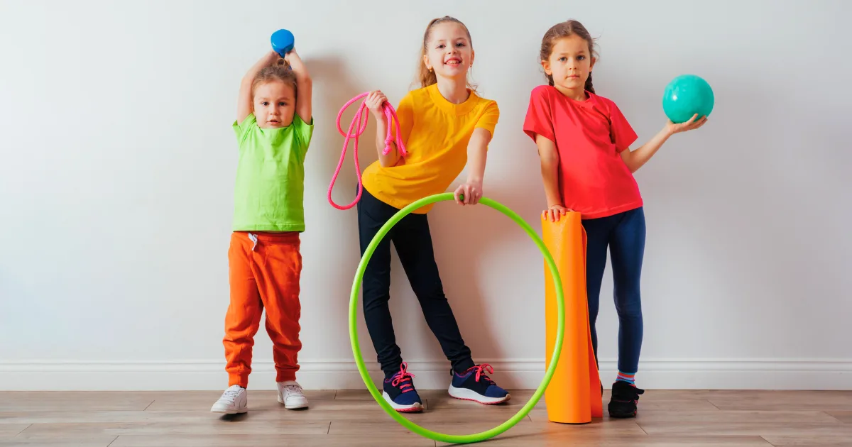 Three children stand indoors against a white wall, each holding sports equipment like a dumbbell, jump rope, and hula hoop—tools that support movement and cognitive development. They wear colorful t-shirts and pants, appearing happy and active.