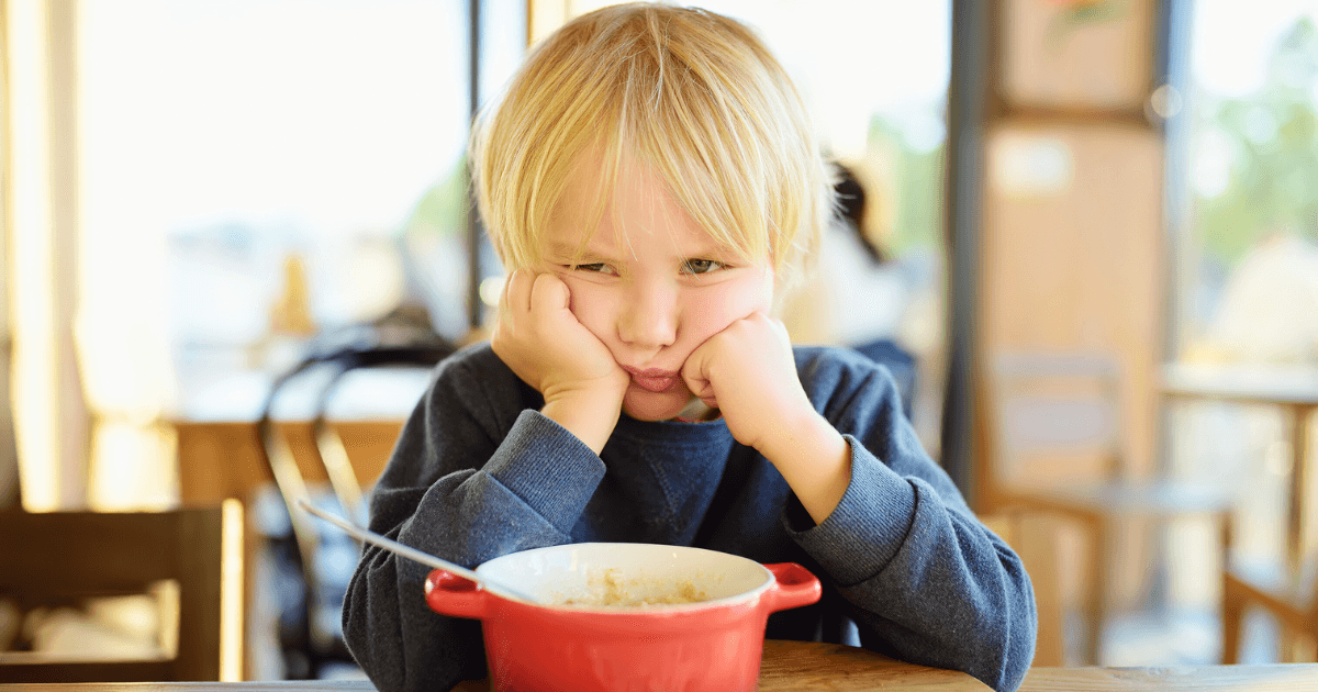 A young child with blond hair rests their face in their hands and looks unhappy while sitting at a table with a red bowl of food, perfectly capturing the struggles many parents face with picky eaters. The background is softly blurred.