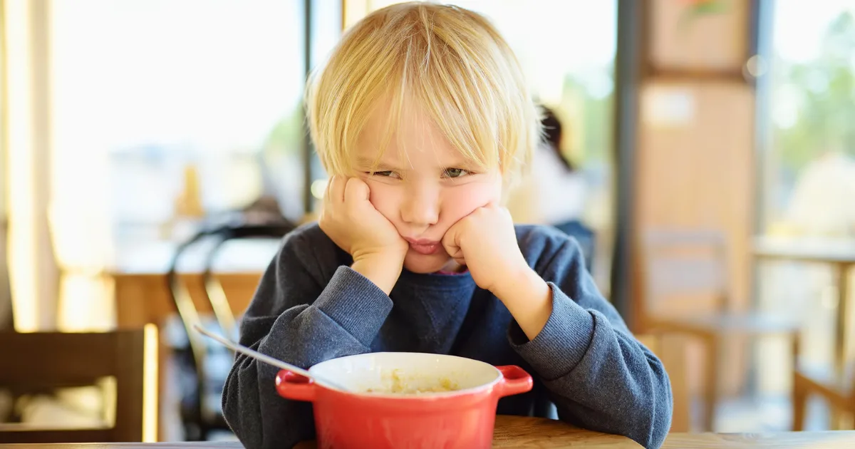A young child with blond hair rests their face in their hands and looks unhappy while sitting at a table with a red bowl of food, perfectly capturing the struggles many parents face with picky eaters. The background is softly blurred.
