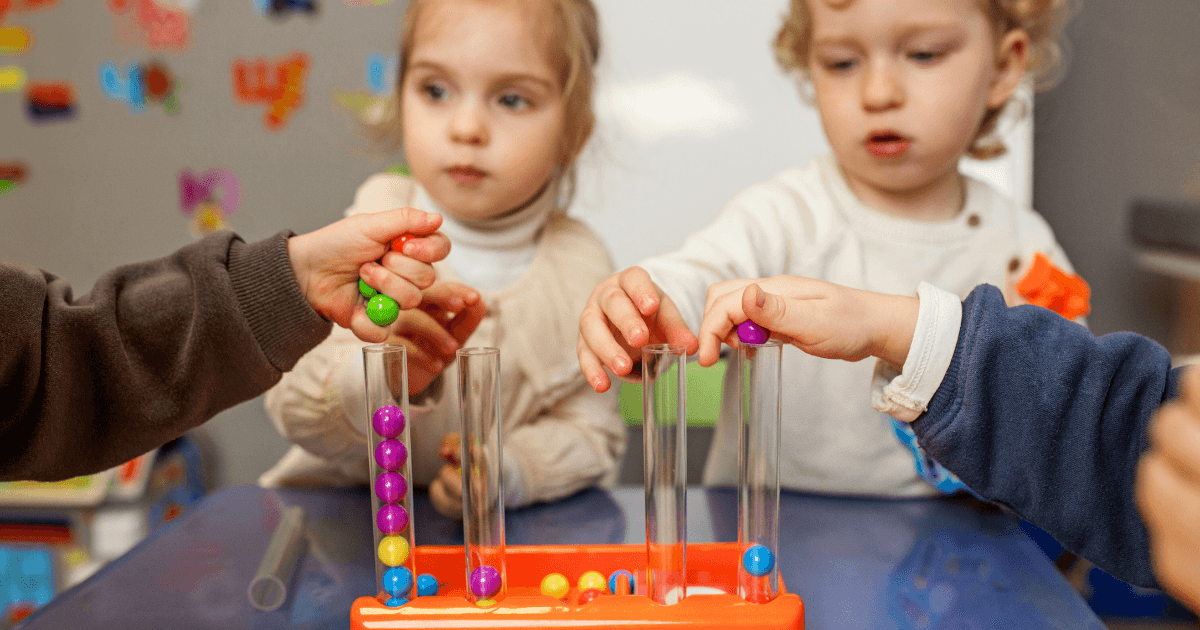 Three young children engage in a play-based activity, placing colorful balls into clear vertical tubes on a table in an academic preschool classroom. Their focus is on the task, with blurred educational materials visible in the background.