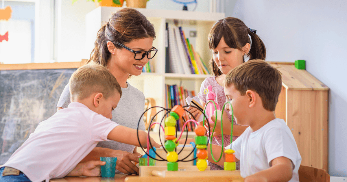 A woman and three young children engage in play-based learning with colorful educational toys at a table in a bright classroom. Shelves with books and other toys are visible in the background.