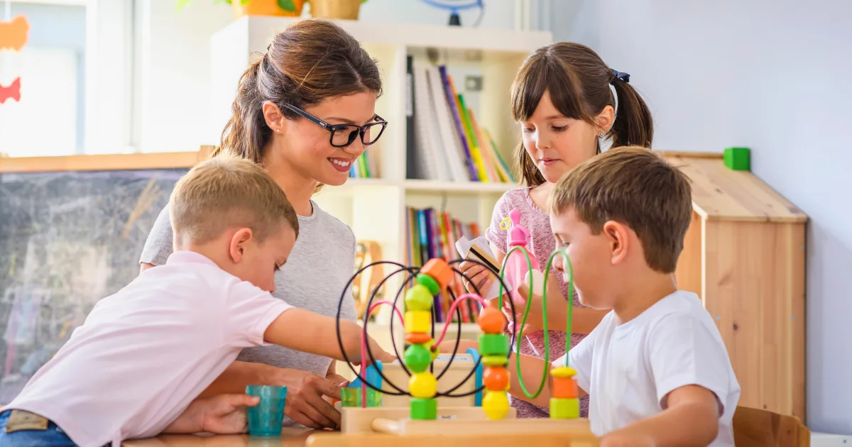 A woman and three young children engage in play-based learning with colorful educational toys at a table in a bright classroom. Shelves with books and other toys are visible in the background.