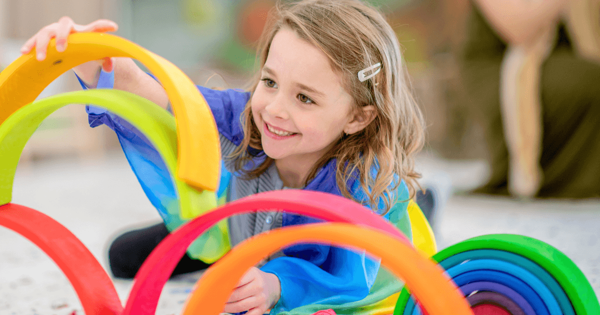 A young girl with light brown hair and a hairclip smiles while playing on the floor with colorful, semi-circular stacking toys at a play school near me, arranged in a cheerful rainbow pattern.