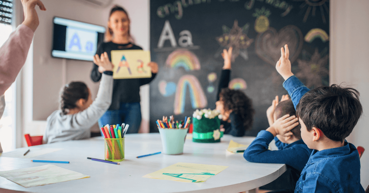 A teacher stands by a chalkboard displaying the letter A to young students during a pre-reading lesson, as some eagerly raise their hands. The classroom desk has colored pencils, paper, and drawings.