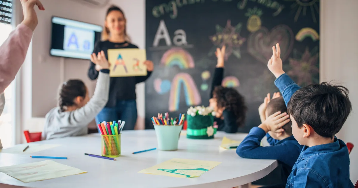 A teacher stands by a chalkboard displaying the letter A to young students during a pre-reading lesson, as some eagerly raise their hands. The classroom desk has colored pencils, paper, and drawings.