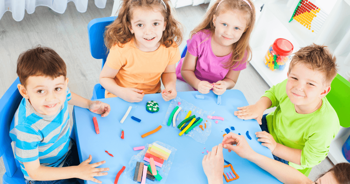 Five young children sit around a blue table, smiling and playing with colorful modeling clay. Various shaped clay pieces and tools are spread on the table, showcasing fun preschool activities in a bright, cheerful classroom.
