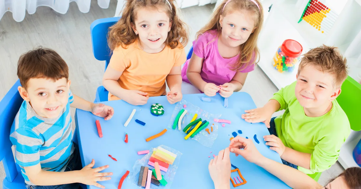 Five young children sit around a blue table, smiling and playing with colorful modeling clay. Various shaped clay pieces and tools are spread on the table, showcasing fun preschool activities in a bright, cheerful classroom.