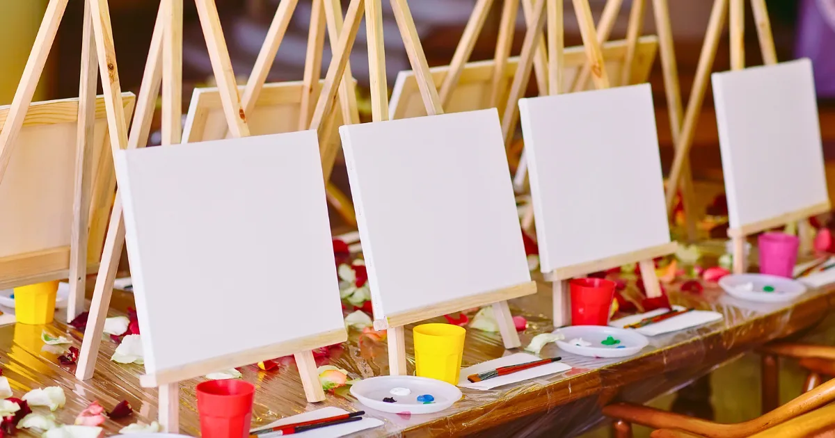 A row of blank canvases on wooden easels is set up with paintbrushes, paint palettes, and colorful cups in an inviting art studio, suggesting a painting class ready to begin.