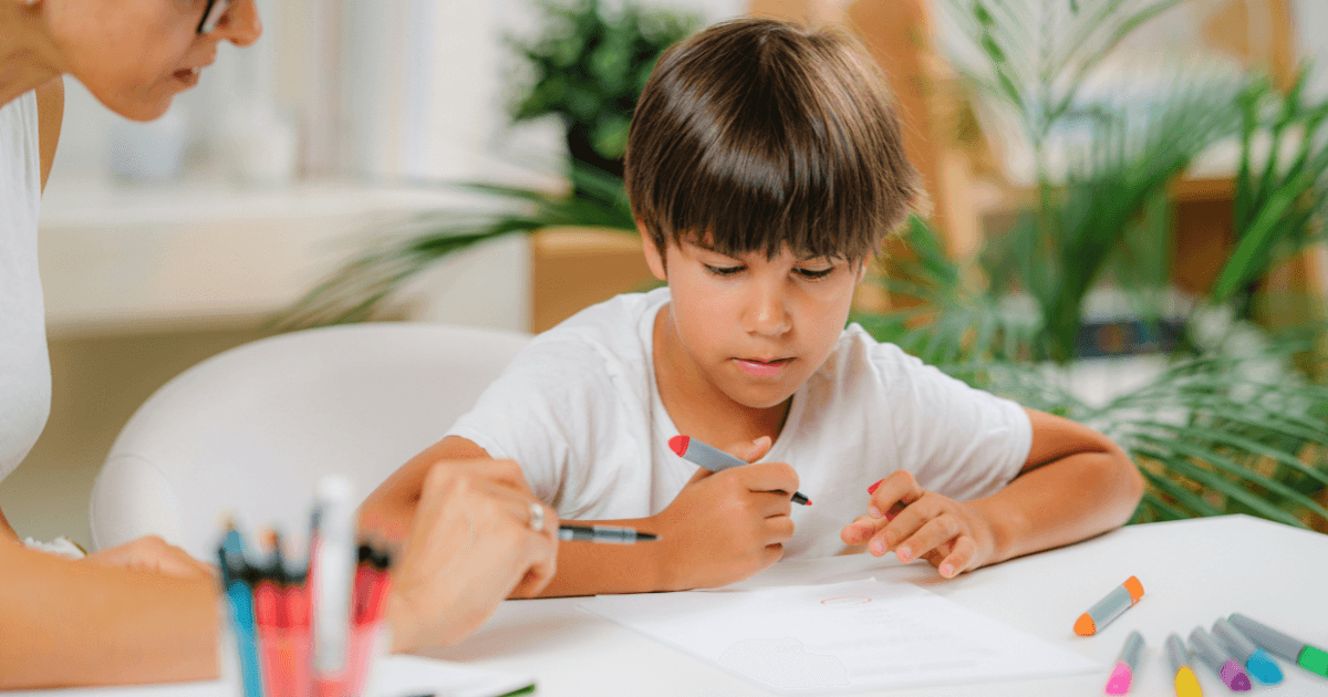 A young boy sits at a white table drawing with colored markers during a preschool assessment, while an adult next to him points at his paper. Various markers are scattered on the table, and green plants are visible in the background.
