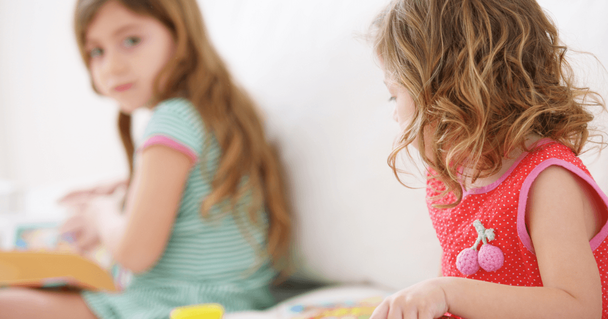 Two young girls with long hair sit indoors. One in a green-striped dress looks forward, while the other in a red dress with a cherry decoration looks at her, demonstrating positive preschool behavior management as they play or read together.
