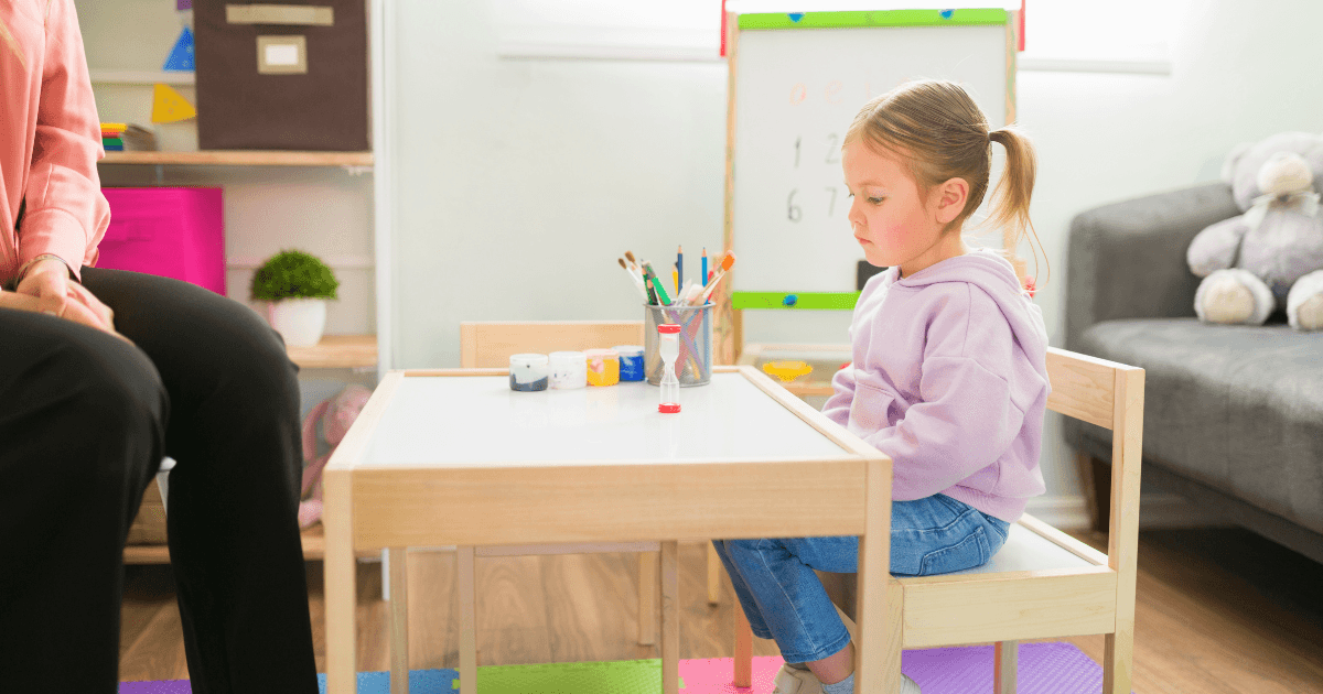 A young girl in a purple hoodie sits thoughtfully at a small table with art supplies in a brightly lit room, while an adult nearby observes her. Toys and a whiteboard hint at activities addressing preschool behavior problems.