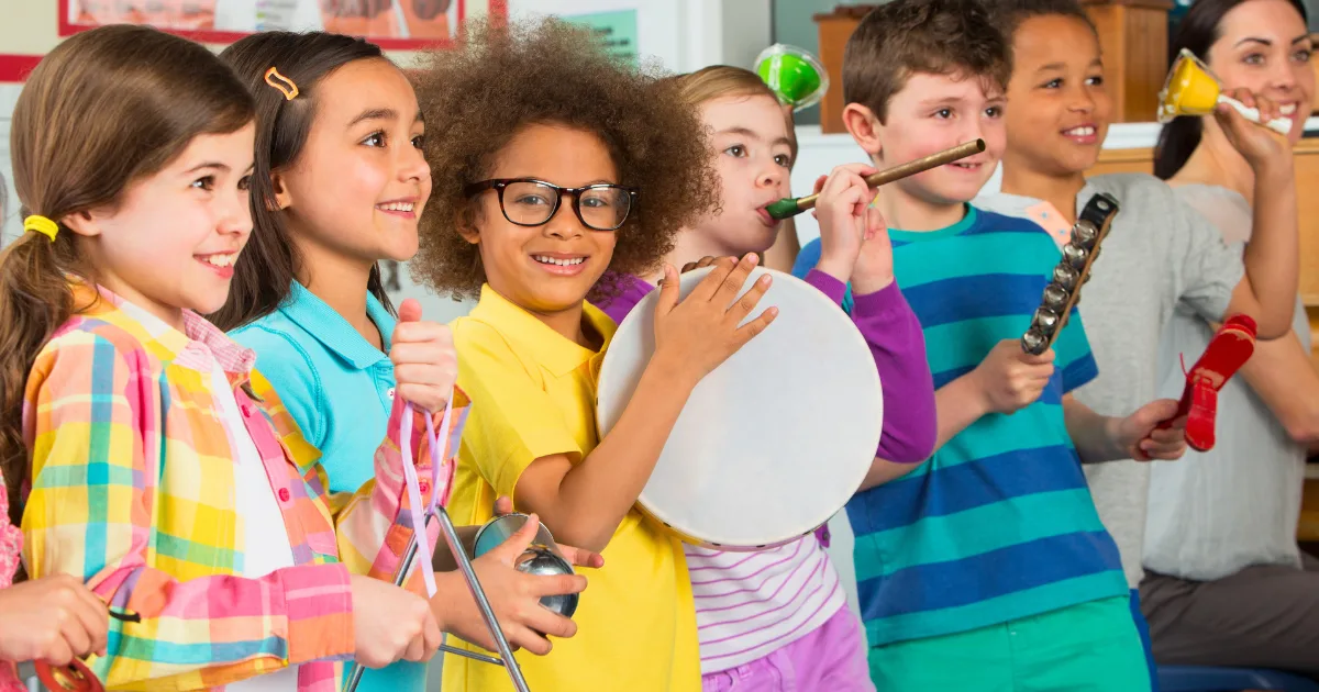 A group of smiling children stand in a row playing various musical instruments, such as maracas, tambourine, and triangles, in a brightly lit classroom during community events.