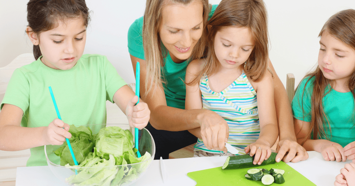 A woman helps a young girl slice cucumber while two other girls, all wearing green tops, prepare lettuce in a bowl. They are sitting together at a table, enjoying a fun cooking activity.