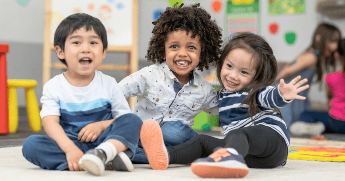 Three young children sit on a classroom floor, smiling and laughing. One child waves, while colorful classroom decorations and curriculum comparison charts are visible in the background.