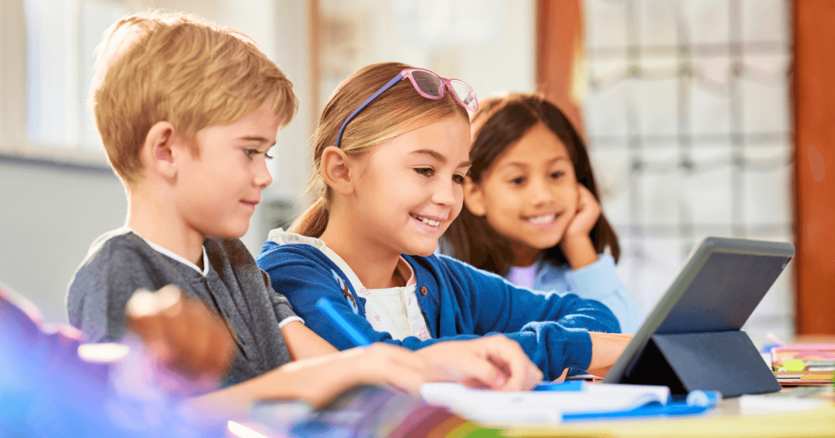 Three children sitting at a desk in a classroom, smiling and looking at a tablet together as they explore their digital portfolios. One girl wears glasses on her head while the others sit beside her, all appearing engaged and happy.