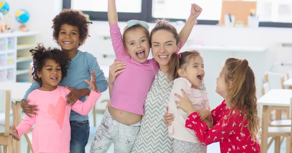 A smiling adult and five young children stand together in a bright classroom, celebrating end-of-year rituals. The children are happy, with some raising their arms or hugging each other and the adult. The scene is cheerful and friendly.