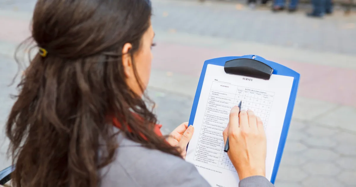 A person with long brown hair holds a clipboard and fills out a family survey form outdoors, using a pen to mark answers on a checklist.