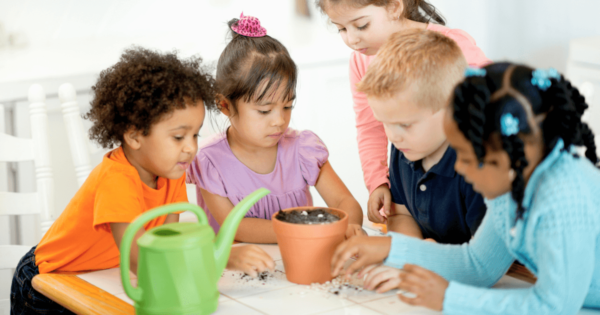 Five young children gather around a table, gardening as they plant seeds in a flowerpot together. A green watering can is nearby, and the kids are focused and engaged in the activity.