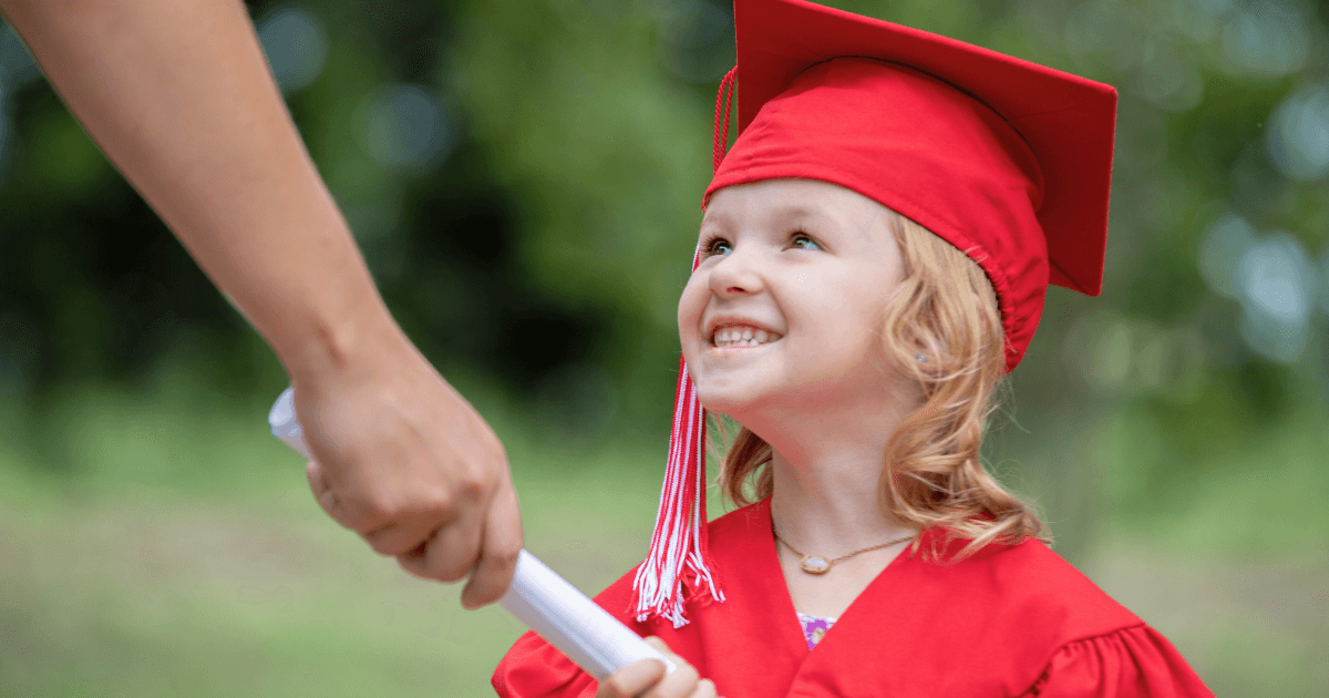 A young child in a red graduation cap and gown smiles brightly during her preschool graduation, looking up at an adult handing her a rolled diploma, with a green, blurred outdoor background.