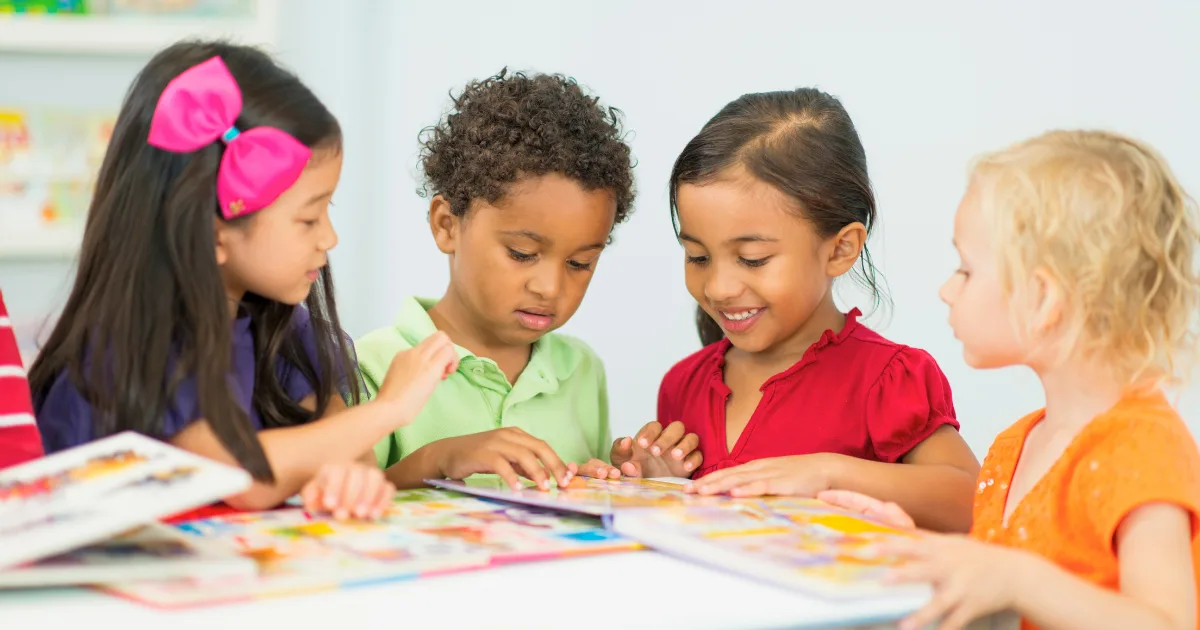 Four young children sit together at a table, looking at colorful picture books and smiling. They appear engaged in preschool learning activities, sharing the books and creating a cheerful, collaborative atmosphere.