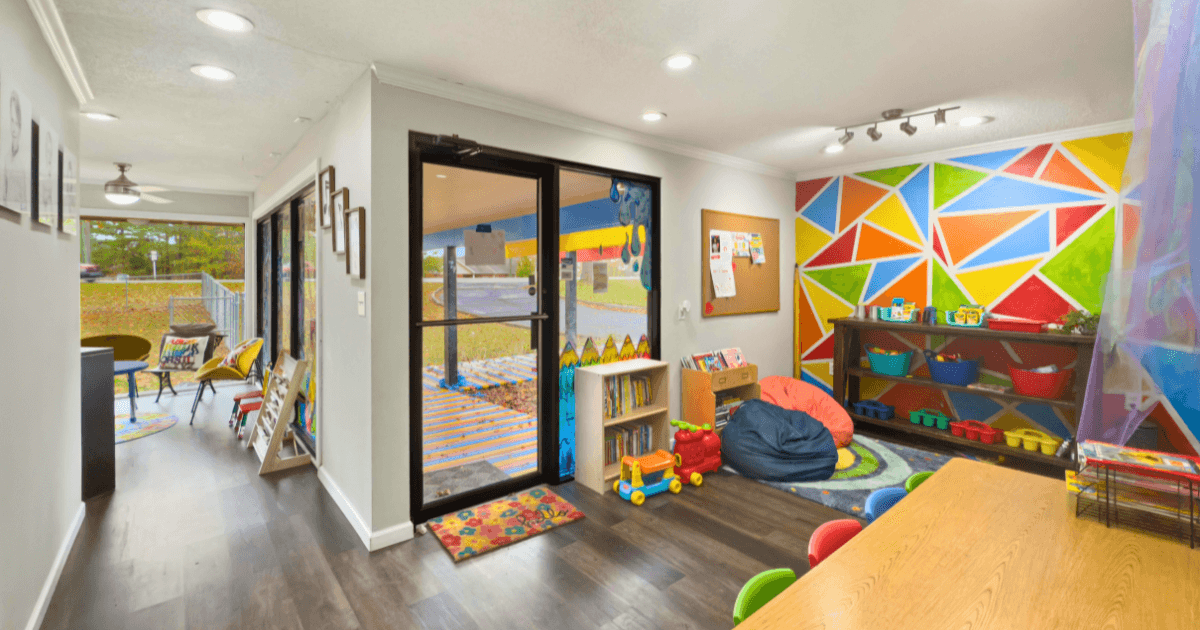 A bright, colorful classroom with a geometric mural, toys, bookshelves, beanbag, and a table with small chairs. Large windows and glass doors let in natural light. Welcoming and organized learning centers await young children.
