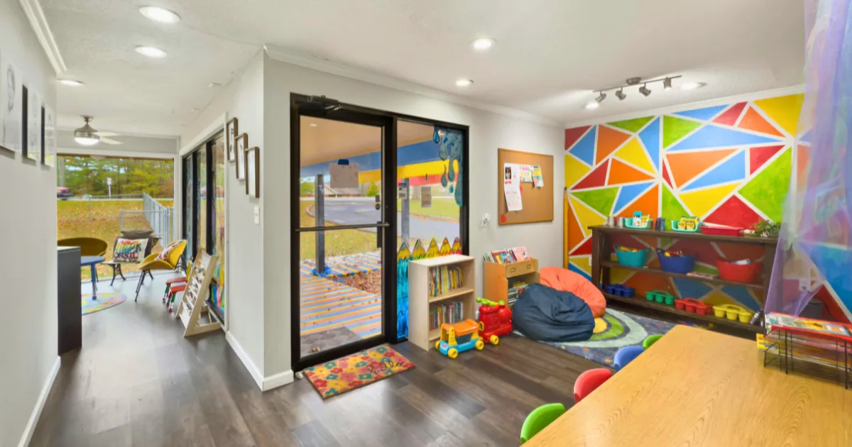 A bright, colorful classroom with a geometric mural, toys, bookshelves, beanbag, and a table with small chairs. Large windows and glass doors let in natural light. Welcoming and organized learning centers await young children.