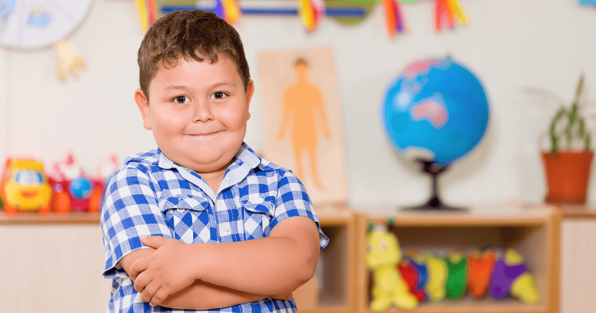 A young boy in a blue and white checkered shirt stands smiling with arms crossed in a colorful classroom, surrounded by toys, a globe, books, and decorations—capturing the joy of reaching preschool learning milestones.