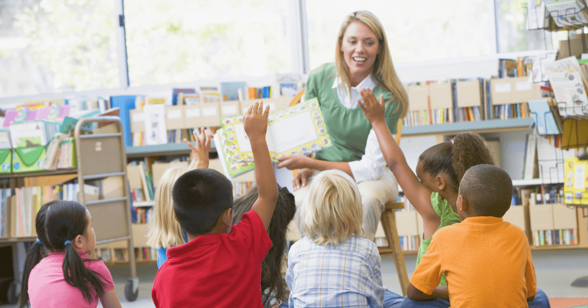 A teacher sits in front of a group of young children in a classroom, smiling and holding a book. Thanks to library partnerships, several children seated on the floor eagerly raise their hands to answer a question.