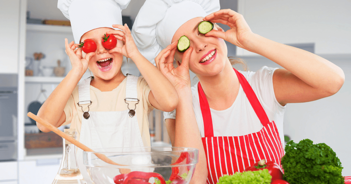 A woman and a child wearing chef hats and aprons playfully hold sliced vegetables over their eyes in the kitchen, surrounded by fresh produce and mixing bowls, smiling and enjoying fun cooking activities together.