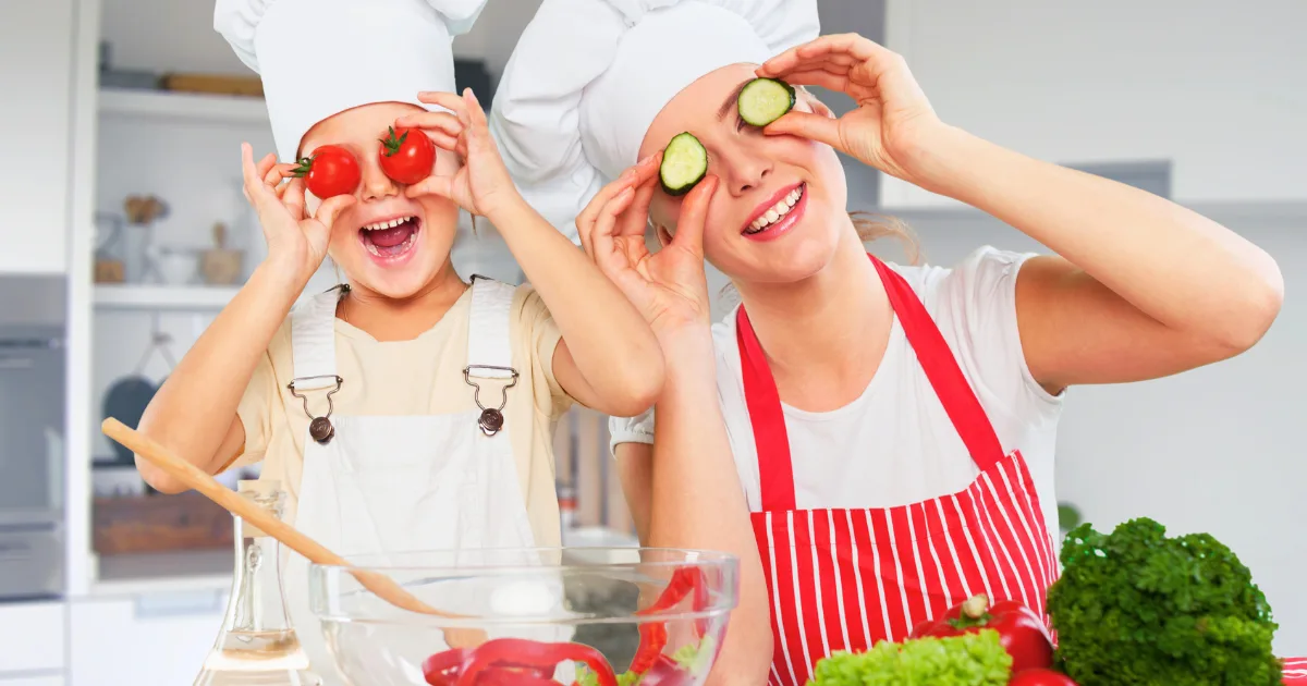 A woman and a child wearing chef hats and aprons playfully hold sliced vegetables over their eyes in the kitchen, surrounded by fresh produce and mixing bowls, smiling and enjoying fun cooking activities together.