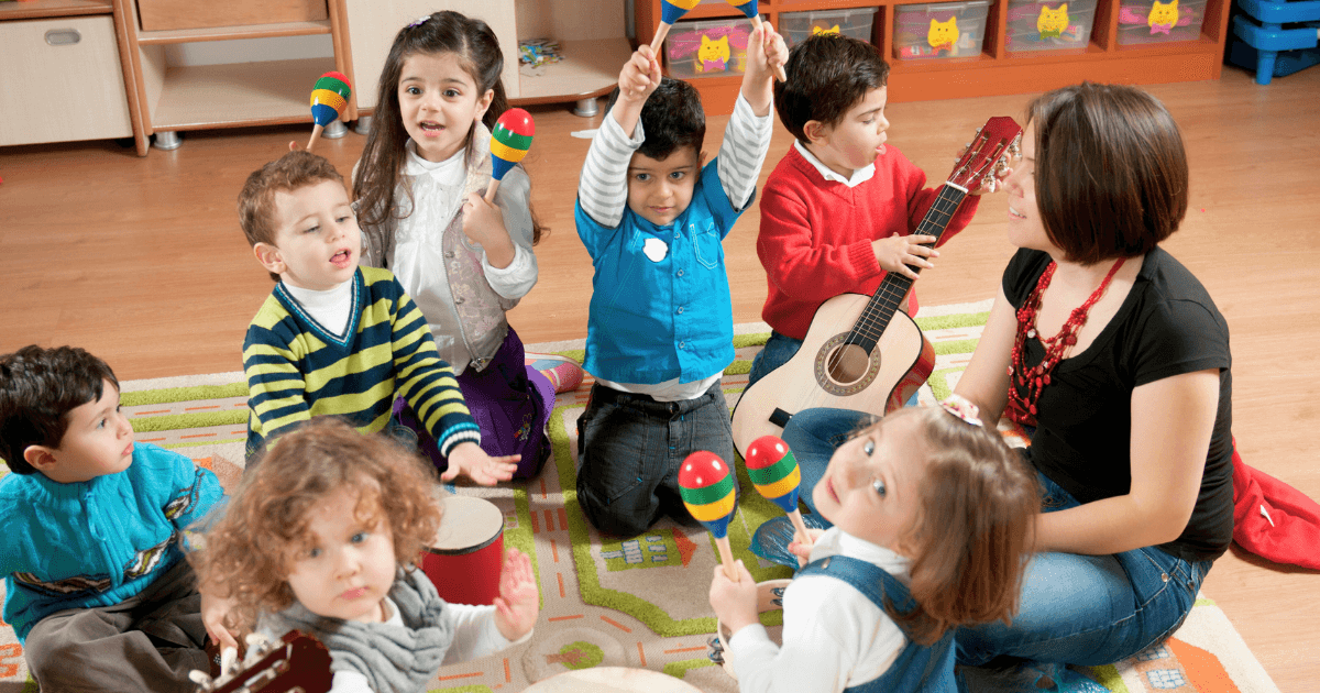 A group of young children sit in a circle on a colorful rug with a teacher, playing musical instruments such as maracas, a drum, and a guitar, enjoying a fun classroom music activity.