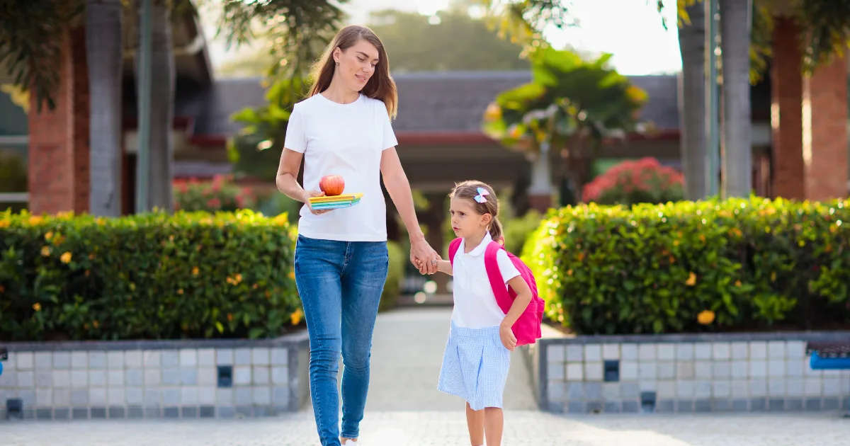 A woman holding books and an apple walks hand in hand with a young girl wearing a backpack, heading to or from school. Sunlight filters through greenery, creating a warm scene perfect for those searching for a preschool near me.